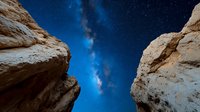 Dramatic night sky view through canyon walls, with bright stars and Milky Way galaxy visible between towering sandstone cliffs under dark blue starry sky.