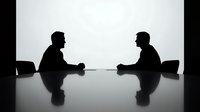 Two business professionals in silhouette sitting across from each other at conference table during meeting or negotiation in modern office setting.