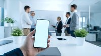 Hand holding smartphone with blank white screen in modern office with blurred business team meeting in background for mobile app mockup presentations.