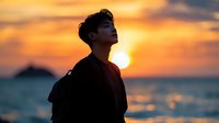 Young Asian male silhouette against dramatic orange sunset sky and ocean horizon, profile view showing contemplative expression and peaceful solitude at dusk.