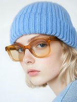 Young woman with blonde hair wearing light blue knit beanie and amber transparent glasses against white background, natural makeup and freckles, casual winter style.