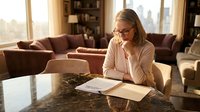Middle-aged Caucasian businesswoman with glasses working from home office, reviewing documents at marble table in modern living room during golden hour.