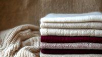 Neatly folded warm knitted sweaters and cardigans in neutral beige and burgundy colors stacked on wooden surface, cozy autumn and winter wardrobe essentials.