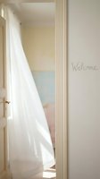 Open white door with sheer curtain flowing in sunlight, Welcome inscription on wall, soft pastel interior, warm inviting home atmosphere.