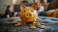 Ceramic piggy bank surrounded by scattered coins on textured surface, shallow depth of field creates soft background blur for financial savings concept.