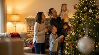Caucasian family of four celebrating holidays together in warmly lit living room decorated with illuminated evergreen tree and festive garland, parents holding children while enjoying festive season at home.