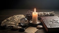 Mystical fortune telling setup with tarot cards, crystal ball, burning candle, sage bundle and ancient symbols on dark wooden table for divination practices.
