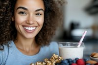 Young mixed-race woman with curly hair smiling while enjoying healthy breakfast with granola, berries and milk smoothie in modern kitchen setting.