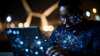 Young African American male professional working on laptop at night with digital network visualization overlay and wind turbine in background. Green energy concept.