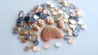 Delicate rose gold paw print surrounded by shimmering metallic sequins and beads scattered on white surface, creating magical pet-themed decorative arrangement.