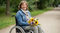 Smiling mature Caucasian woman in wheelchair holding colorful sunflower bouquet outdoors in park setting for disability awareness and inclusion concepts.