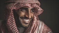 Portrait of Middle Eastern man wearing traditional red and white keffiyeh headdress, smiling warmly against dark background.