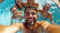 Diverse group of young friends taking selfie while swimming in pool, capturing summer fun and joy. Bearded man holds camera with smiling women in background.