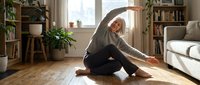 Senior Caucasian woman stretching on living room floor, doing yoga side bend, smiling, promoting active aging and healthy lifestyle at home.