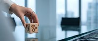 Professional hand placing wooden cube with gear mechanism icon on modern glass desk in bright office environment for business strategy concepts.