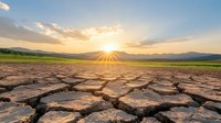 Cracked dry soil in desert with dramatic sunset rays breaking through clouds over mountain landscape. Drought concept showing climate change effects.