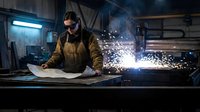 Male welder in protective gear working with metal in industrial workshop, creating sparks during welding process for manufacturing and construction projects.