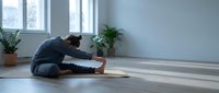 Young woman practicing yoga seated forward bend pose on mat in bright modern room with plants and natural light streaming through windows.