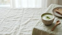 Creamy soup with herb garnish served in rustic bowl on linen tablecloth with bread and wooden spoon for food styling and culinary content.