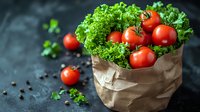 Fresh red tomatoes and crisp green lettuce leaves in brown paper bag on dark background with scattered peppercorns, moody food photography composition.