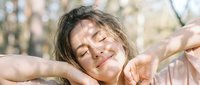 Young woman with dark hair and fair complexion enjoying outdoor sunlight with eyes closed and arms stretched, expressing joy and relaxation in nature.