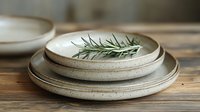 Handmade ceramic dinnerware set with natural speckled glaze and fresh rosemary herb garnish on rustic wooden table for organic dining and kitchen decor.