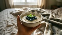 Overhead view of hand holding creamy oatmeal bowl topped with fresh blueberries and mint leaf, styled on cozy bed with natural window light for breakfast and wellness content.