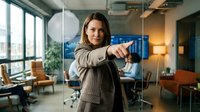 Professional businesswoman pointing forward in modern office space with confident gesture and warm smile during corporate meeting or presentation.