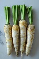Fresh white daikon radishes with bright green tops arranged in row on marble background, showing natural texture and organic imperfections of root vegetables.