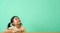 Young Asian female child wearing yellow shirt gazing upward thoughtfully against mint green background, suitable for educational, inspirational, and childhood concept designs.