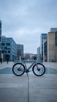 Modern fixed-gear bicycle parked on urban plaza with contemporary architecture and blurred pedestrians in background. Suitable for cycling, transportation, and city lifestyle content.