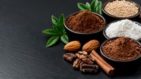 Raw ingredients for healthy baking including cocoa powder, flour, nuts and spices arranged on dark slate background with fresh basil leaves and copy space.
