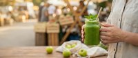 Woman holding fresh green smoothie drink in glass jar at outdoor farmers market with organic vegetables and healthy lifestyle concept.
