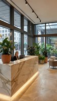 Modern office lobby with marble reception desk, blonde businesswoman receptionist, large windows, indoor plants, contemporary lighting and urban city view.
