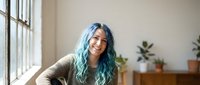 Young woman with vibrant blue-green hair smiling confidently in bright modern interior with large windows and plants for lifestyle branding content.