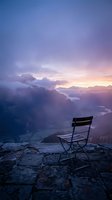Empty chair positioned on rocky mountain cliff overlooking misty valley at sunrise, with dramatic purple and pink sky, golden light breaking through clouds, and distant landscape below for contemplative themes.