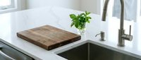 Wooden cutting board with fresh green herbs on white kitchen countertop next to stainless steel sink and faucet for cooking and food preparation.