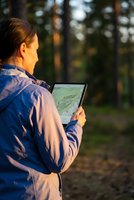 Young Caucasian woman hiker using digital tablet with map application in forest during golden hour sunset for outdoor navigation and adventure planning.