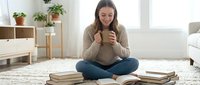 Young pregnant woman enjoying hot beverage while reading books in cozy modern living room with plants and natural light for relaxation and wellness.