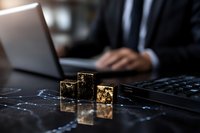 Gold metallic cubes on dark reflective surface with businessman working on laptop in background, creating modern business atmosphere for finance concepts.