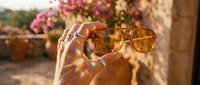 Young woman with medium skin tone holding trendy amber-tinted sunglasses outdoors in sunny garden setting with blooming flowers and natural light for lifestyle and fashion use.