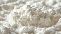 Fine white flour powder macro shot showing soft texture and delicate particles scattered across surface, creating gentle shadows and highlights in natural light.