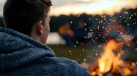 Young man in hoodie watching campfire sparks rise into twilight sky during outdoor evening gathering with warm glowing embers and peaceful atmosphere.