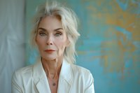 Elegant mature woman with platinum blonde hair and striking green eyes wearing white blazer against turquoise and gold wall background, perfect for beauty, fashion or business concepts.