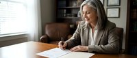 Senior woman with gray hair and light skin tone, wearing professional business attire, signing documents at wooden desk in office setting. Suitable for corporate, legal, and business concepts.