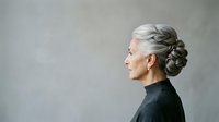 Senior woman with gray hair styled in a bun, wearing black turtleneck, profile view against neutral gray background. Mature adult portrait.