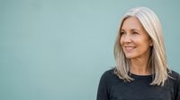 Mature woman with long platinum blonde hair smiling against soft blue background, suitable for healthcare, wellness, lifestyle and corporate marketing materials.