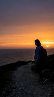 Woman hiker with backpack standing on coastal cliff path at golden sunset overlooking ocean, travel and adventure lifestyle photography.