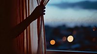 Hand pulling back curtain revealing blurred city lights at dusk through window creating warm intimate atmosphere for home decor and lifestyle concepts.