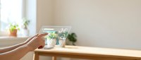 Hands holding transparent tablet displaying holographic interface icons in bright modern interior with wooden desk and plants for futuristic technology concepts.